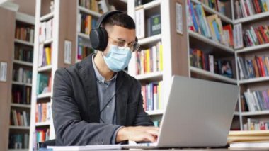 Maskeli adam dizüstü bilgisayar kullanıyor ve kütüphanede kulaklıkla ses dinliyor.
