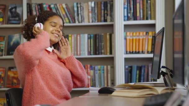 Черная девушка в библиотеке слушает музыку в наушниках — стоковое видео