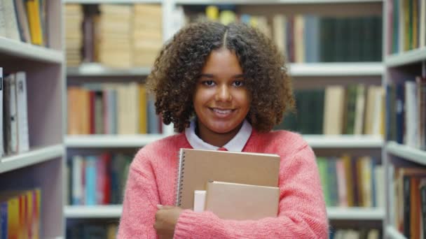 Estudiante afroamericana posando con libros en la biblioteca universitaria — Vídeos de Stock