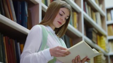 Uzun saçlı, beyaz gömlekli ve yelekli, kitaplığa yaslanmış ve sesli kitap okuyan güzel bir kadın. Düşük açılı üniversite öğrencisi kütüphanede edebiyatın tadını çıkarıyor. Boş zaman kavramı.
