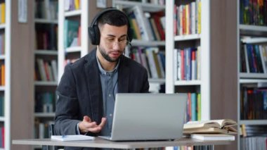 Gözlüklü neşeli adam video görüşmesi için laptop ve kulaklık kullanıyor, webcam ile konuşuyor, el kol hareketi yapıyor, telekonferans veya zoom etkinliği yapıyor, kütüphanede çevrimiçi eğitim alıyor, arka planda kitaplıklar var.