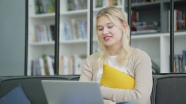 Sarı saçlı neşeli genç kız koltukta oturuyor, yastığa sarılıyor ve bilgisayarla video görüşmesi yapıyor, web kamerası kullanıyor, internette iletişim kuruyor. Teknoloji kavramı