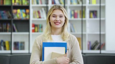 Uzun sarı saçlı, sıradan kıyafetler giyen, elinde kitaplarla kameraya bakan, arka planda bulanık kitaplıklar olan mutlu bir kız. Üniversite kütüphanesindeki kız öğrencinin portresi. Eğitim kavramı