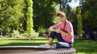 Gündelik kıyafetli genç bir kadın yaz parkında bankta oturuyor ve yanındaki akıllı telefon, dizüstü bilgisayar ve kitaplarla sosyal medyada geziniyor. Öğrencilerin dışarıda rahatlarken parallaks fotoğrafı.