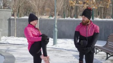 Pembe ve siyah spor kıyafetleri giyen arkadaşlar kış parkında birlikte egzersiz yapıyorlar, bacaklarını esnetiyorlar, yumruk tokuşturuyorlar ve koşmaya başlıyorlar. Açık hava aktivitesi ve sağlıklı yaşam tarzı. Spor anlayışı