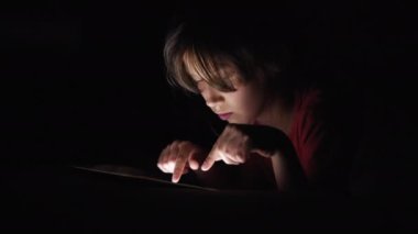 Çocuk karanlıkta dokunmatik tablet ekranına dokunuyor, geceleri online oyun oynuyor, internette geziniyor, gülümsüyor ve eğleniyor, ekrandan yüzüne ışık geliyor. Teknoloji kavramı