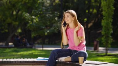 Uzun saçlı, pembe tişörtlü ve kot pantolonlu bir kadın yaz parkında bankta oturuyor ve yanında telefon, karton bardak ve dizüstü bilgisayarla konuşuyor. Teknoloji kavramı