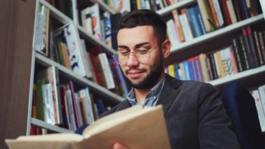 Cheerful young man with eyeglasses sitting by bookshelves and reading book, flipping pages. Low angle portrait of reader enjoying literature in book shop or library. Concept of education