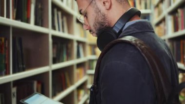 Young bearded man with eyeglasses having backpack on shoulder and headphones on neck holding tablet and searching for certain books in library or in book shop. Concept of education