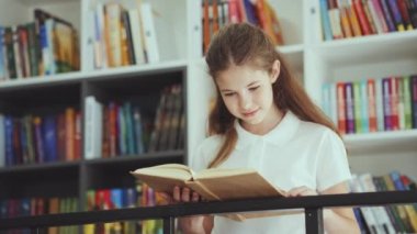 Smiling child standing with book by bookshelves and reading. Portrait of smart girl wearing white polo shirt enjoying literature in book shop or school library. Concept of education