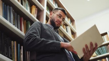 Young bearded man with eyeglasses standing next to bookshelves and reading book, flipping pages. Low angle portrait of intelligent reader enjoying literature in book shop or library