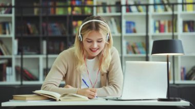 Happy teenager wearing headphones sitting at desk and writing in notebook, laptop in front of her, student doing assignment and listening to music. Concept of technology and education