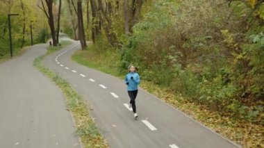 Slow motion fit female jogger running in park in the morning, autumn leaves lying on ground, male jogger running in the opposite direction, tracking shot from above. Healthy lifestyle