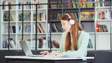 Akıllı kız kütüphanede kitap yığınının önünde oturuyor ve dizüstü bilgisayarla ödev yapıyor, kulaklıkla müzik dinliyor ve ritme göre hareket ediyor, arka planda kitaplıklar var. Eğitim kavramı