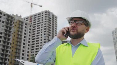Sakallı müteahhit, kasket ve güvenlik yeleği giyiyor inşaat alanında yürüyor ve telefonla konuşuyor, çok katlı binalar ve bulanık arka planda vinçler var. Vurulan adamın izini sürüyorum.