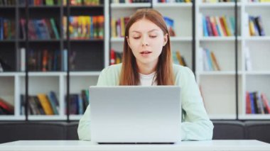 Kütüphanede dizüstü bilgisayarında video görüşmesi yapan, webcam 'le konuşan telekonferans ya da zoom etkinliği olan, arka planda bulanık kitaplıklar olan genç bir kız. Online rota. Uzaklık öğrenimi