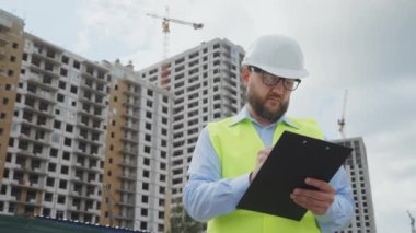 Miğfer ve güvenlik yeleği giyen sakallı adam dosya klasörü tutuyor ve inşaat halindeki çok katlı binalara karşı notlar tutuyor. Bölgede çalışan düşük açılı müfettiş kaliteyi kontrol ediyor.