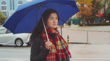 Kareli atkı takan çekici genç bir kadın yağmur altında cadde kavşağının önünde mavi bir şemsiyeyle bekliyor. Arabalar ve arka plandaki insanlar bulanık. Şehir hayatı kavramı