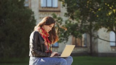 Deri ceket giyen, eşarplı ve kot pantolonlu genç bir kadın güneşli parkta bankta oturuyor ve dizüstü bilgisayarla çalışıyor, bina geçmişi bulanık. Öğrenci dışarıda çalışıyor. Eğitim kavramı