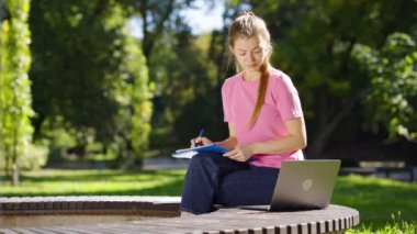 At kuyruklu, pembe tişörtlü ve kot pantolonlu genç bir kız öğrenci yaz parkında bankta oturuyor ve ödev yapıyor, kağıda not alıyor, yanında duran dizüstü bilgisayarını kullanıyor. Eğitim kavramı