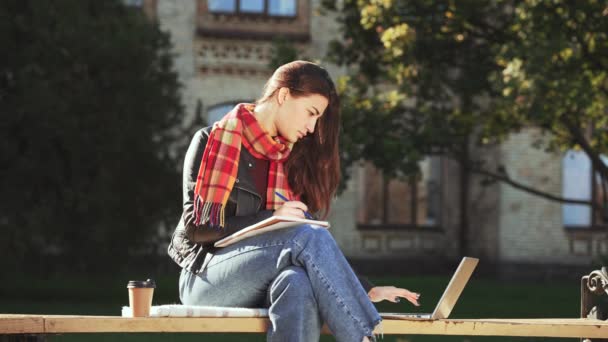 年轻的女性穿着皮夹克 格子围巾和牛仔裤坐在长椅上 在纸上做笔记 看着旁边的笔记本电脑 在网上学习 在外面学习 — 图库视频影像