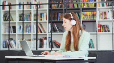 Kütüphanede çalışan, müzik dinleyen, dizüstü bilgisayarla çalışan, şarkı söyleyen, kameraya bakan, arka planda bulanık kitap rafları olan beyaz kız öğrenci. Eğitim kavramı