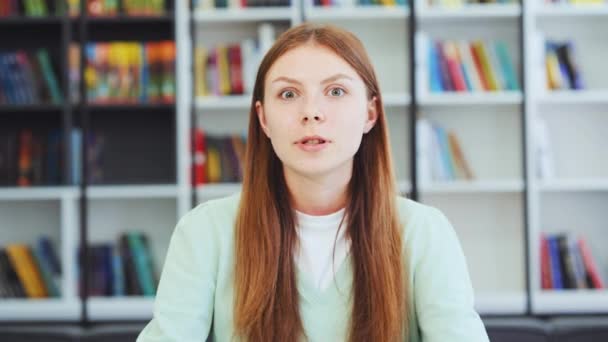 在网上会议上 有一头长发的多愁善感的年轻女学生对着网络摄像头说话 摆出姿势 模糊了书架的背景 为表达观点的年轻女性准备的衣服 交流的概念 — 图库视频影像