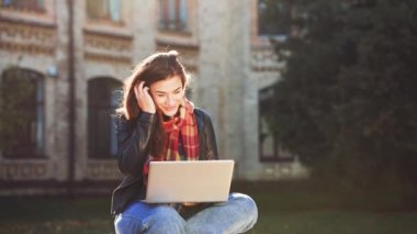 Deri ceket giyen ve eşarp takan genç bir kadın gün batımında dizüstü bilgisayarla görüntülü görüşme yapıyor. Kampüs binasının geçmişi bulanık. Çevrimiçi iletişim kavramı