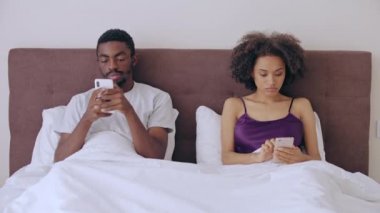 Genç siyah çift yatakta oturuyor, akıllı telefonları kaydırıyor, birbirlerini görmezden geliyorlar.