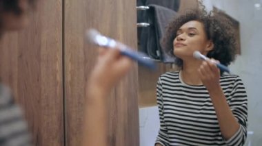 Güzel siyah kadın makyaj yapıyor banyoda fırçayla gülümsüyor, güzellik bakımı yapıyor.