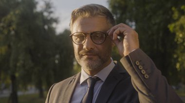 Kendine güvenen orta yaşlı bir adam kameraya bakıyor, gözlükleri ayarlıyor, yüksek kalifiye profesyonel.