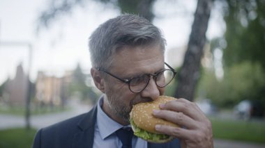 40 'larında bir iş adamı parkta hamburger yiyor, sağlıksız bir yaşam tarzı, fast food.