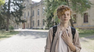 Kızıl saçlı okul çocuğu okul bahçesinde tek başına dikiliyor, diğer çocuklarla sozileşme sorunu yaşıyor.