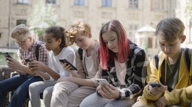 Çok ırklı okul çocukları akıllı telefonlar ve alet bağımlılığıyla bankta oturuyorlar.