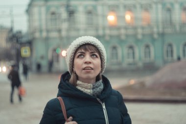 Kışlık şapkalı bir kız şehirde dolaşıp binalara bakıyor.