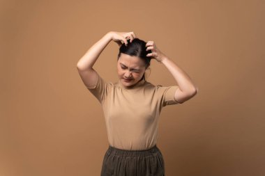 Asyalı kadın sıkılmış ve canı sıkkın, başını kaşıyor, bej arka planda tek başına duruyor..