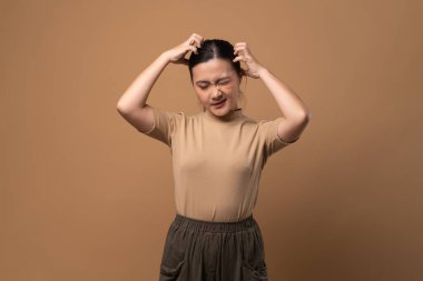 Asyalı kadın sıkılmış ve canı sıkkın, başını kaşıyor, bej arka planda tek başına duruyor..