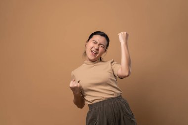 Asyalı kadın kendine güveni tam, yumruğunu göstererek bej arka planda izole bir şekilde zafer jesti yapıyor..