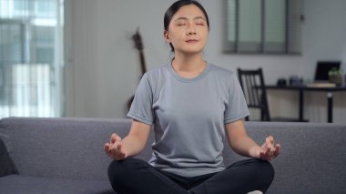 Asyalı kadın sakinleşiyor, derin derin nefes alıyor yoga jestinde ellerini tutuyor evdeki oturma odasında kanepede oturuyor.. 