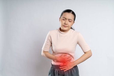 Asian woman was sick with stomach ache, putting her hands on belly with red spot, standing isolated on white background.