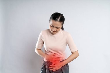 Asian woman was sick with stomach ache, putting her hands on belly with red spot, standing isolated on white background.