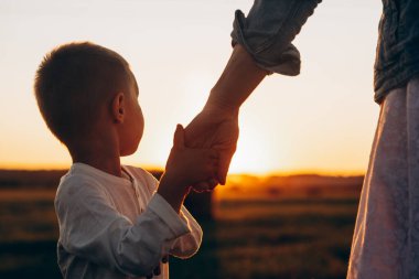 Anne, yaz aylarında doğada çocuğuna rehberlik ediyor. Güven, aile, koruma ve yardım kavramı. Kadın ve çocuğun elleri yaz tarlasının günbatımına yakın.