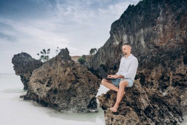 Genç adam sahildeki kayanın üzerinde dizüstü bilgisayar kullanıyor. Rahatlama Tatili Açık Sahil Konsepti