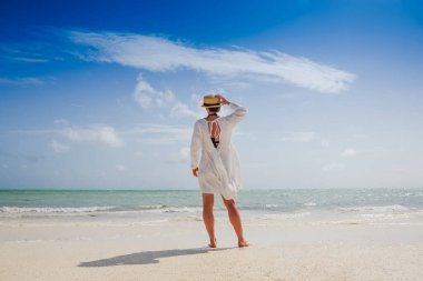 Beyaz elbiseli ve şapkalı mutlu gezgin kadın tropik plaj tatilinin tadını çıkarıyor.