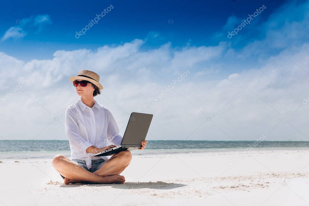 A woman using and typing on laptop computer keyboard while sitting on a beautiful tropical beach. Freelance work, vacations, distance work, social distancing, e-learning, connection, creative professional, new business, meeting online concept