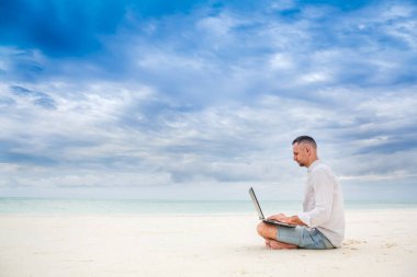 Adam kumsalda mavi denizin önünde dizüstü bilgisayarda yazı yazıyor. Uzaktan çalışmayı kabul et, serbest çalış.