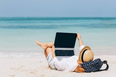 Kumsalda dizüstü bilgisayar kullanan genç bir kadın çalışıyor. Serbest çalışma, tatiller, mesafe çalışması, sosyal uzaklık, e-öğrenme, bağlantı, yaratıcı meslek, yeni iş, online konsept buluşması