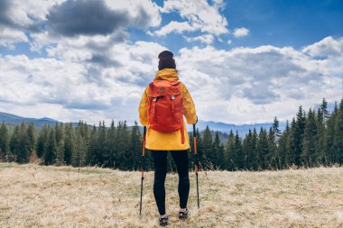Dağlara karşı dağ sırtının yamacında duran mutlu kadın yürüyüşçü, arka planda mavi bulutlu gökyüzü. Kadın mutlu ve manzaraya bakıyor. Seyahat ve aktif yaşam tarzı kavramı.