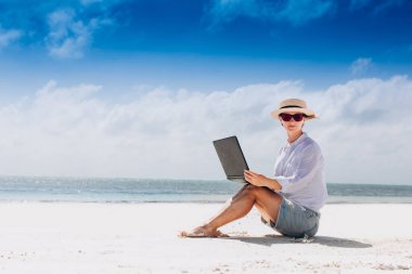 Güzel bir tropik plajda otururken dizüstü bilgisayarda klavye kullanan ve daktilo kullanan bir kadın.