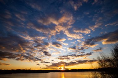 Önünde ağaç siluetleri olan güzel mavi bir göl dramatik bir günbatımı gökyüzünün altında.